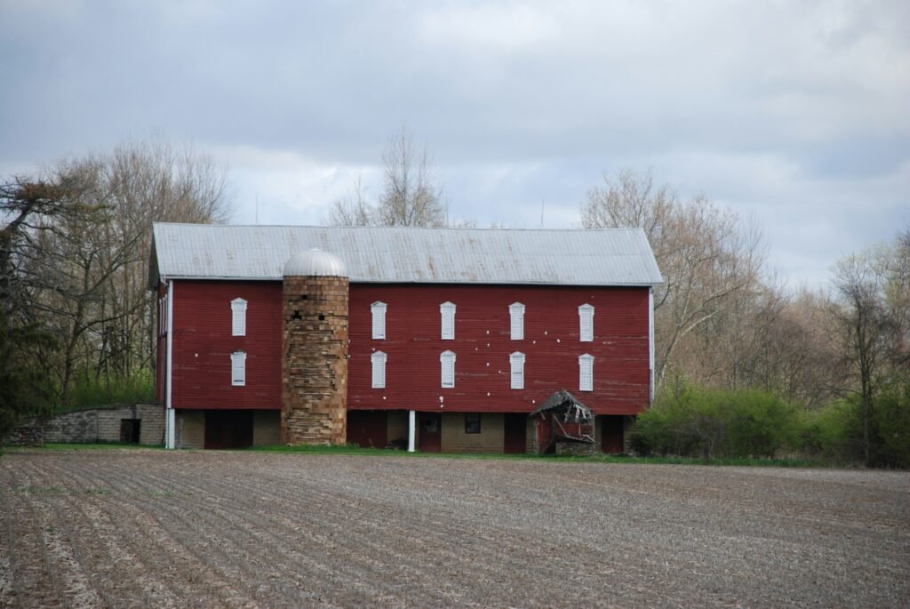 Close-up of a traditional red wooden barn with a central silo, standing beside an open field with bare soil and leafless trees under an overcast sky.